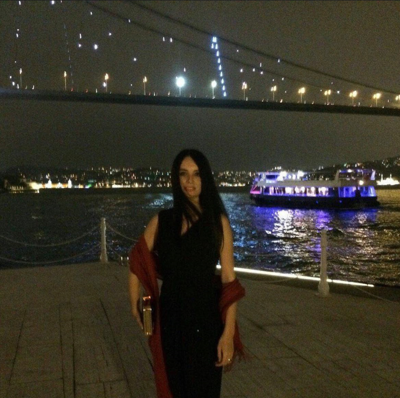 Eda Ketenci, gece İstanbul Boğazı kıyısında, ışıklandırılmış Boğaz Köprüsü ve denizdeki tekneler önünde.